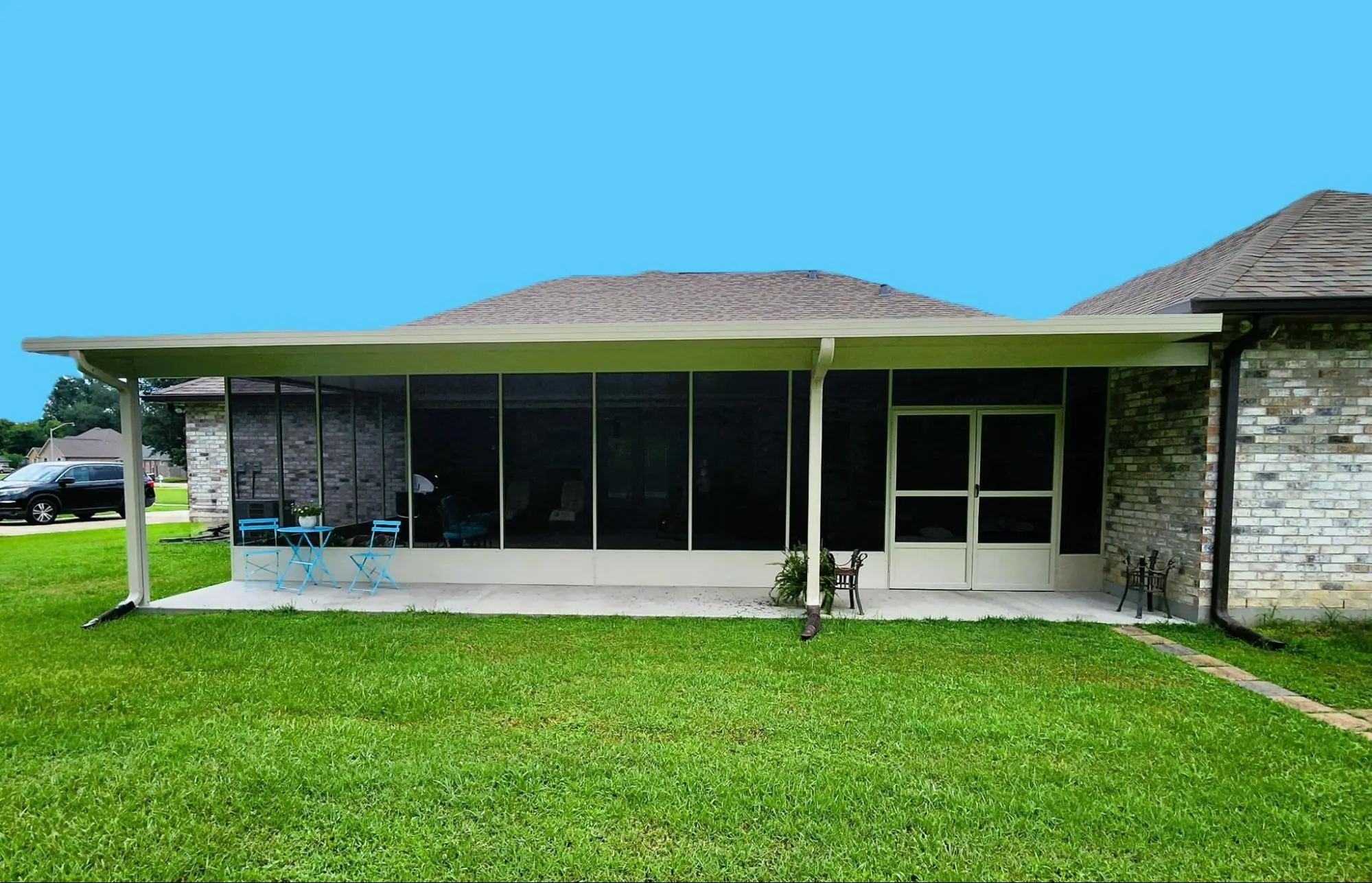 A brick house with a screened-in porch. The porch has a table set with four blue chairs and overlooks a green lawn. The roof is sloped.